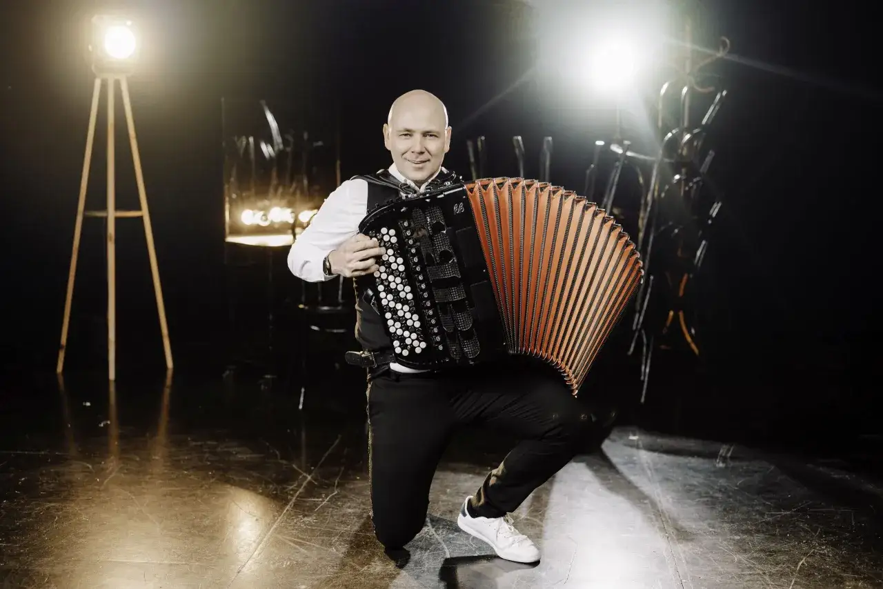 Mężczyzna na klęczkach gra na akordeonie. W tle reflektory i scenografia. Piękna gra na akordeonie.
