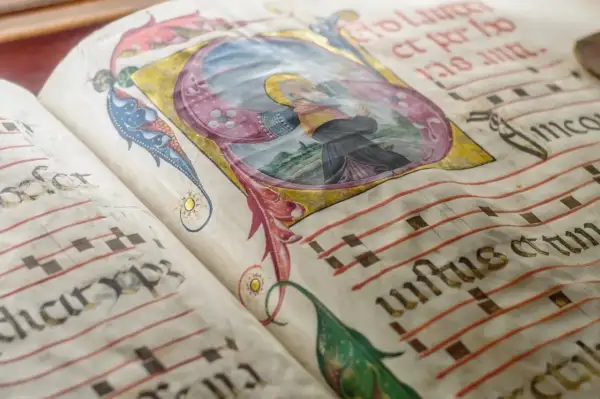 Stara księga z iluminacjami. Widać fragment rękopisu z nutami i ozdobnym inicjałem. Cała nuta jest tu częścią średniowiecznej notacji muzycznej.